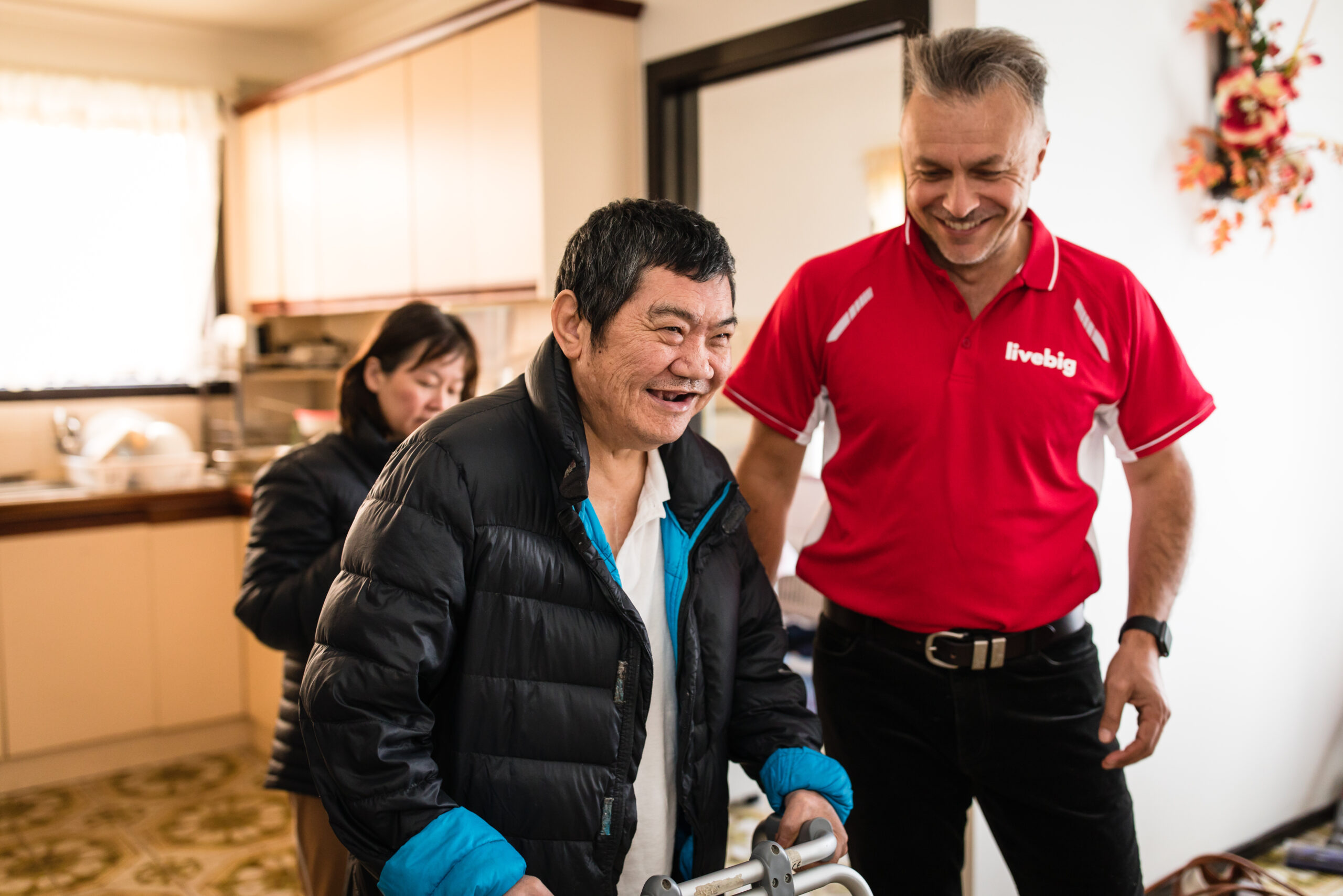 A LiveBig therapist supporting a smiling client using a walking frame at home