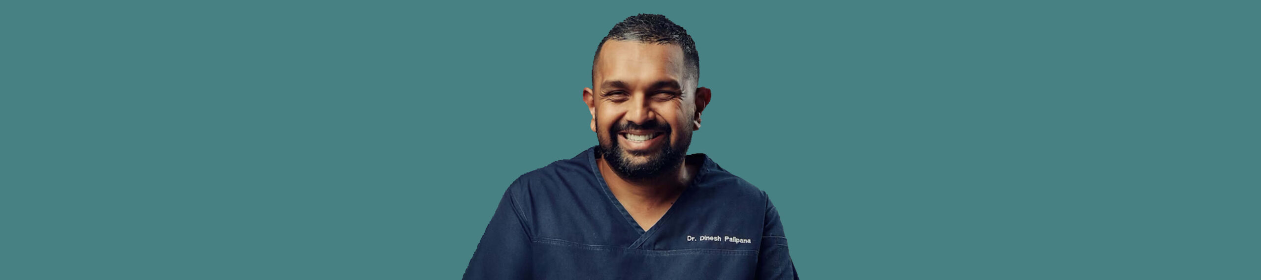 Dr. Dinesh Palipana smiling while wearing blue medical scrubs