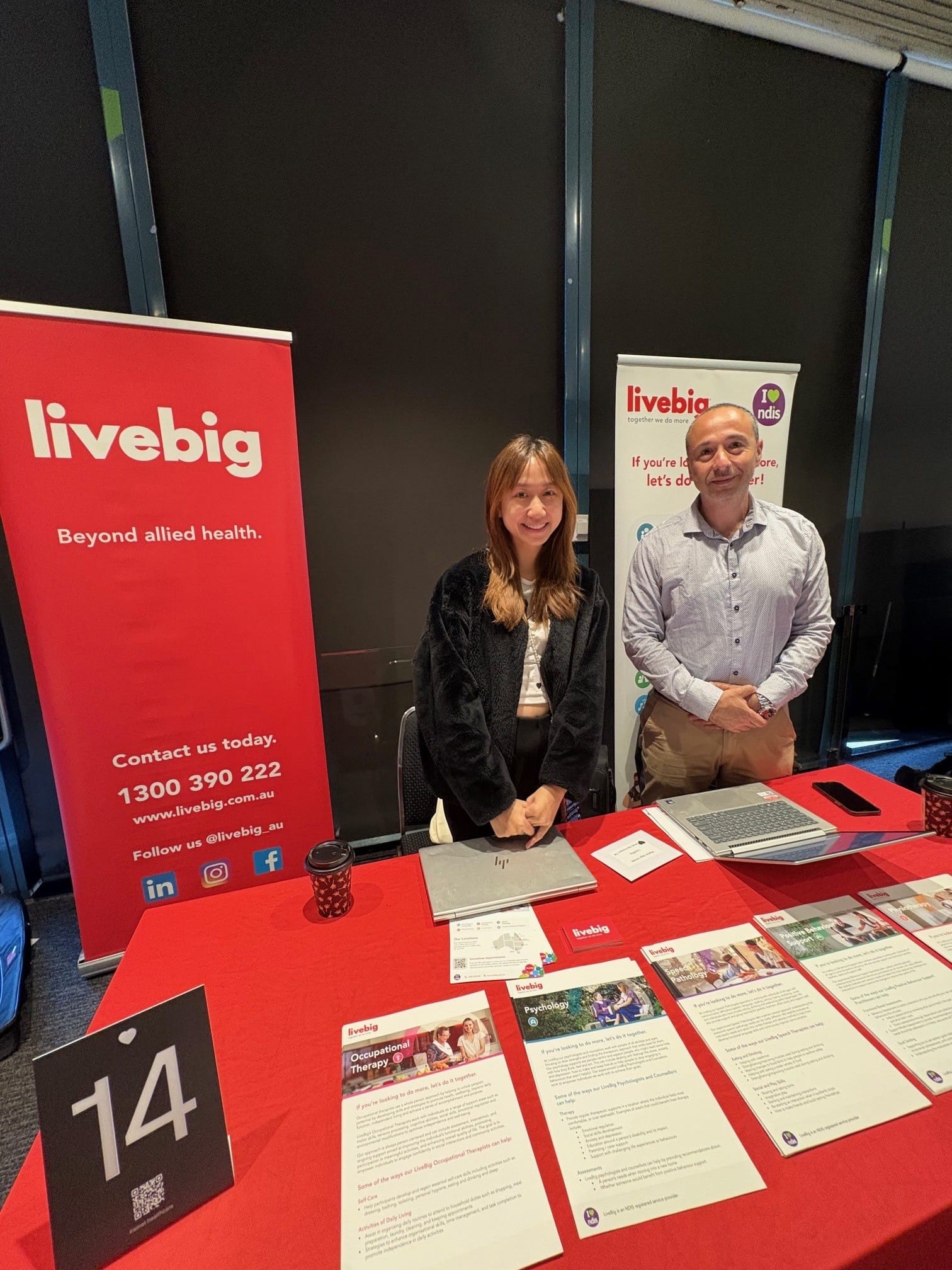 Two LiveBig team members standing behind a promotional booth displaying allied health service brochures