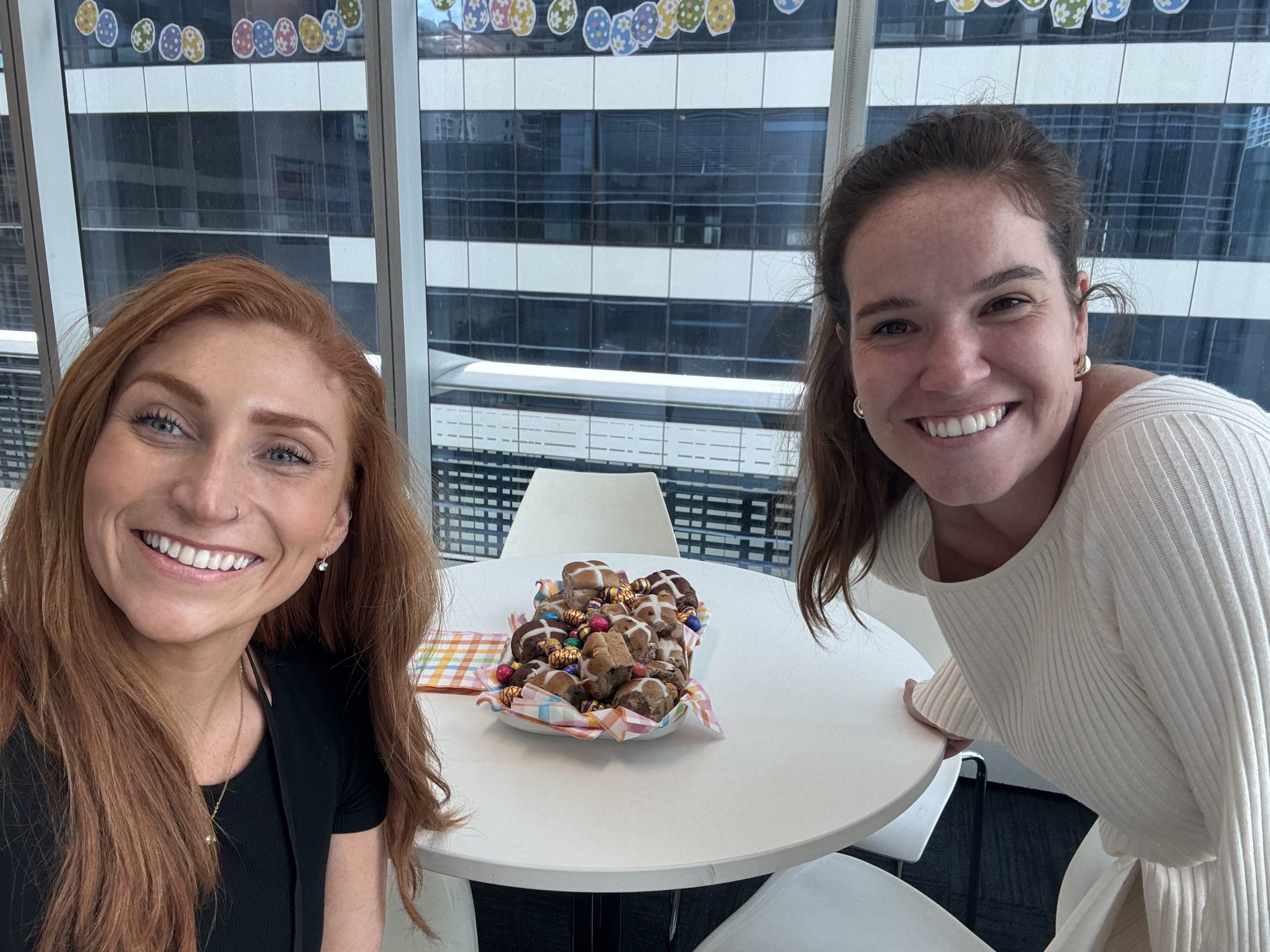 Two Arriba Group employees smiling for a selfie during an Easter workplace celebration with hot cross buns