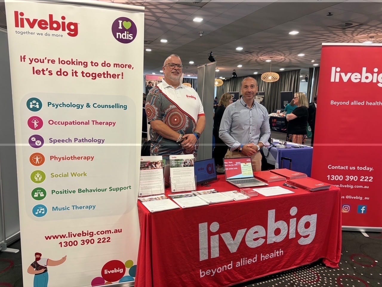Two LiveBig representatives standing behind a branded exhibition booth promoting allied health services
